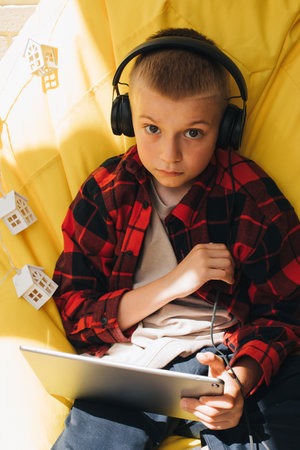 A school-age boy in black headphones sits on a yellow bean bag with a tablet in his hands. Children and gadgets. Front viewの写真素材