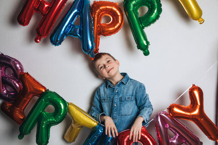 Childrens birthday celebration. A joyful boy, a schoolboy, in a blue shirt against the background of colorful balloons. Front viewの写真素材