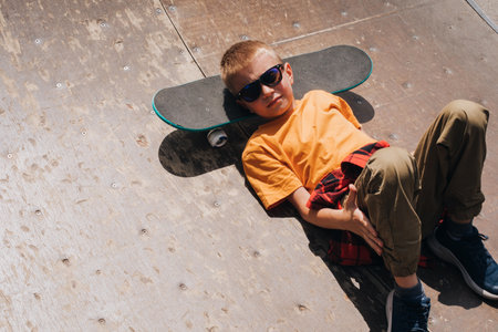 Outdoor activities for children. A cheerful young boy in sunglasses lies on a skateboard on the playground. Caucasian schoolboy on a walk. Top viewの写真素材