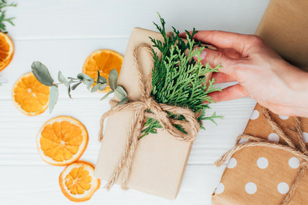 The gift is wrapped by female hands in brown paper and tied with twine with thuja and eucalyptus on a white wooden background. Top viewの写真素材