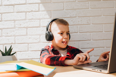 A school-age boy in black headphones sits at a table with a laptop. Children and gadgets. Front view