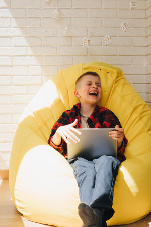 A school-age boy sits on a yellow bean bag with a tablet in his hands. Children and gadgets. Front viewの写真素材