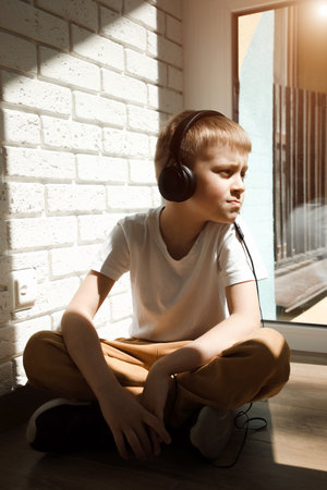 A boy of 6-7 years old in a white T-shirt listens to music in black headphones, sings and looks out...
