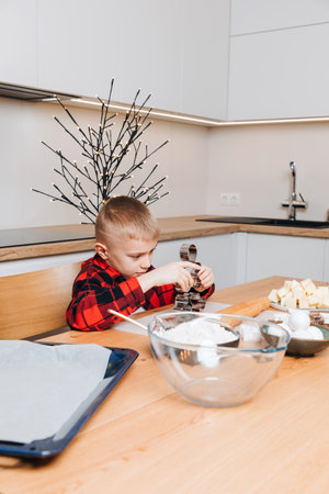 A boy in a red plaid shirt at a wooden table in the kitchen is going to make cookies holding a cookie cutter. Front viewの写真素材