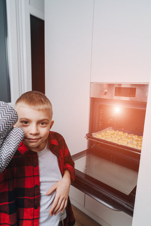 A boy in a red shirt puts a baking sheet with Christmas cookies in the oven. Preparing for Christmas. Cooking cookies at home. Front viewの写真素材