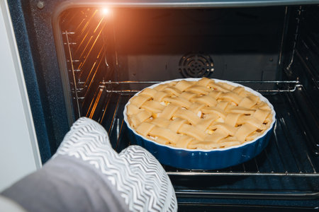 Making desserts at home. Hand in gray thermo mittens put the apple pie in the oven for baking. Front viewの写真素材