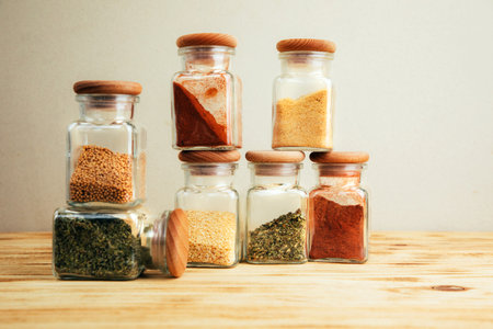 A group of seasonings in glass jars on a light wooden background. Paprika, herbs, mustard, garlic, front view, selective focusの写真素材