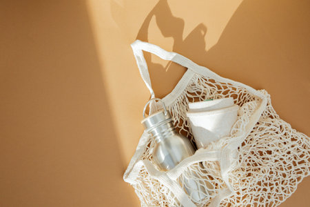 Beige mesh bag for storage with tin bottle and cup for coffee on a brown background. Eco bags for multiple use, conscious consumption. Flat layの写真素材