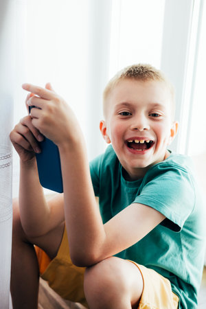 Cheerful school-age boy in a green T-shirt sitting at the window with smartphone. Children and devices. Front viewの写真素材