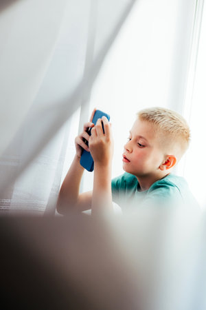 Cheerful school-age boy in a green T-shirt sitting at the window with smartphone. Children and devices. Front viewの写真素材