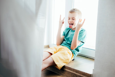 Cheerful school-age boy in a green T-shirt sitting at the window at home. Front viewの写真素材