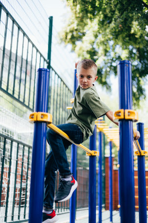 A school-age boy in a green T-shirt spends time on horizontal bars, a playground in sunny weather. Front viewの写真素材