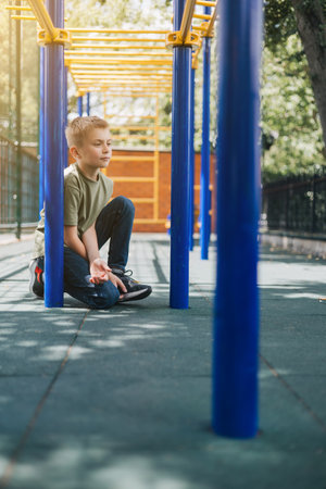 A school-age boy in a green T-shirt spends time on horizontal bars, a playground in sunny weather. Front viewの写真素材