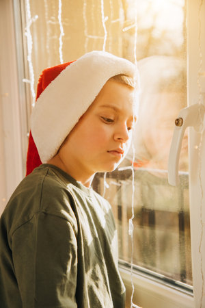 A boy in red Christmas hat near the window in bright sunlight. Children and pets. Portrait of a child. Christmas eve. Front viewの写真素材