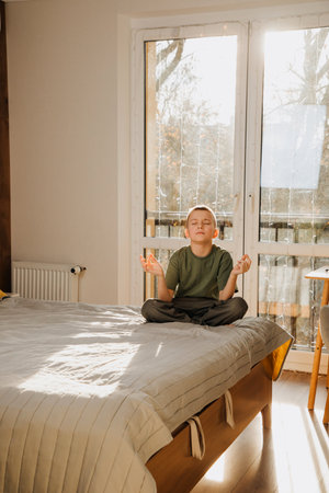 A boy in a green T-shirt meditates on the bed in bright sunlight. Calming with meditation. Portrait of a child. Front viewの写真素材