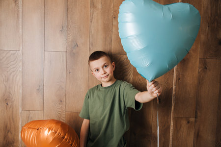A boy in a green T-shirt with an orange and blue balloons in the shape of a heart against a wooden wall. Valentines Day. Portrait of a child. Front viewの写真素材