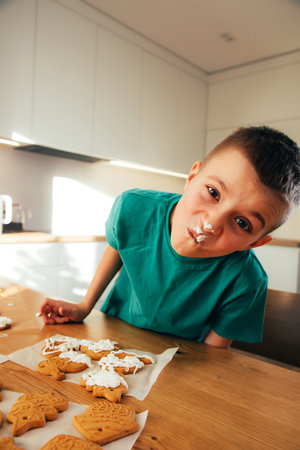 School-age boy in sunny kitchen decorating cookies with white protein icing. Children are helpers in the kitchen. Time spent together with childrenの写真素材