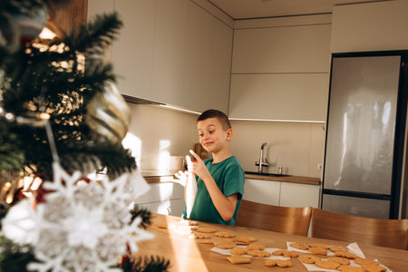 A school-age boy in the kitchen decorates Christmas cookies in sunny weather. Children in the kitchen, homemade pastries. Front viewの写真素材
