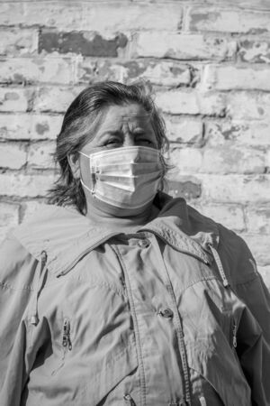 Senior woman stands in a medical mask on her face, which protects against coronavirus and other viruses and diseases on a background of old brick wall. Coronavirus pandemic. Black and white toneの写真素材