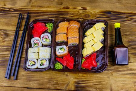 Set of sushi rolls in plastic boxes, soy sauce and chopsticks on wooden table. Top view. Sushi for take away or delivery of sushi in plastic containersの写真素材