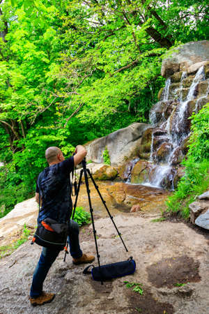 Travel photographer man with professional camera on tripod taking photos of waterfallの写真素材