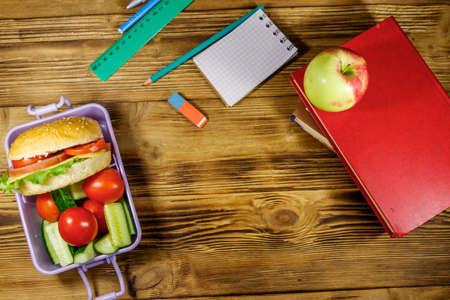 Back to school concept. School supplies, books, apple and lunch box with burgers and fresh vegetables on a wooden table. Top viewの写真素材