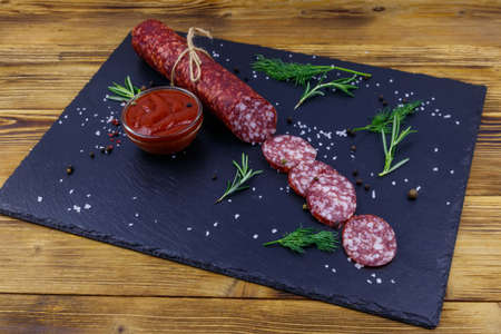 Salami sausage with ketchup and spices on black slate board on a wooden tableの写真素材