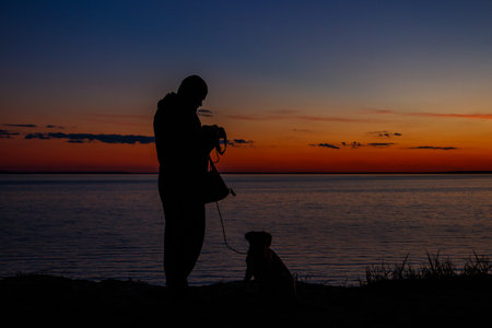 Silhouette of a guy with photo camera and dog on a river shore at sunsetの写真素材