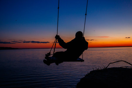 Silhouette of a man on rope swing above river at sunsetの写真素材