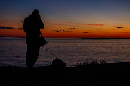 Silhouette of a guy with photo camera and dog on a river shore at sunsetの写真素材