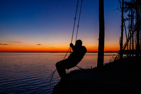 Silhouette of a man on rope swing above river at sunsetの写真素材