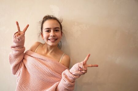 Portrait of pretty positive happy teenage girl gesturing peace dressed in pink knitted loose sweater. Toned photo with copy space on background of grunge beige wallの写真素材