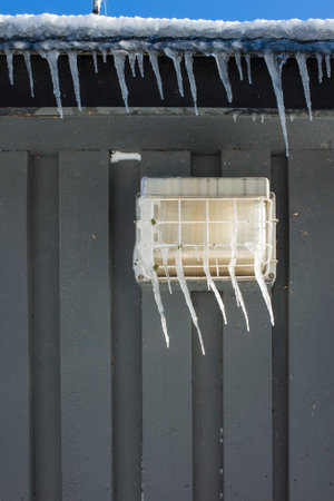 Icicles hang from roof and street lamp on gray painted wooden wall close up abstract photo, Cold...