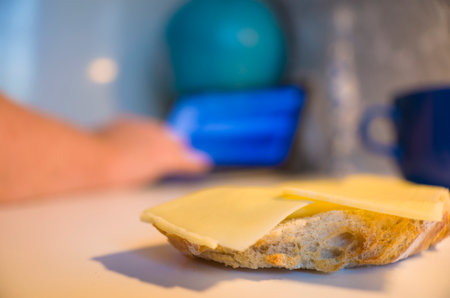 Cheese sandwich close up photo with selective focus, blurred backdrop of white table with hands typing on laptop, blue mug, Working from home, freelance, self employment, Snack timeの写真素材