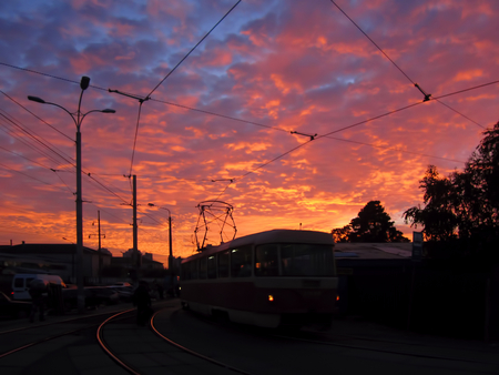Transport trolley, sundown time. Dark street lanterns, tram with tramway on the background of yellow orange blue sky with red clouds. Sundown today, time of sunset, sunrise and sunset, streetcar.の写真素材