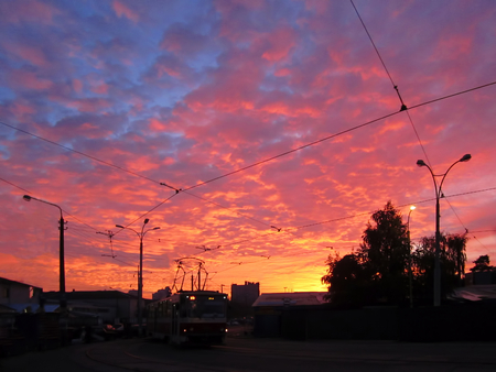 Sunset tomorrow. Black street lights, tram on tramway on the background of yellow orange blue sky with light clouds. Sundown today, time of sunset, transport trolley, sunrise tomorrow.の写真素材
