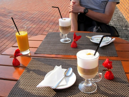 Table with two glasses of cappuccino, orange juice, three candies in red wrapper,  three black tubes. Guy sits at the table, one can see men arms, hands, fingers, pinkie ring. Friends, togetherの写真素材
