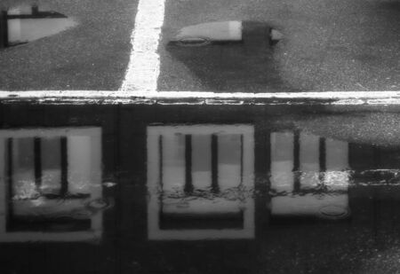Window reflection in the road asphalt puddle. Rain drops in the puddle. Three windows. Black and white. Raining spring weather. Dark flooded house property. Water reflection house in the puddle.の写真素材
