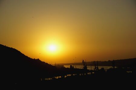 Panoramic view to yellow sunset sun sits on hill and Black sea bay. Cranes stand near shore on water. Comfortable port in Inkerman, Sevastopol, Crimea, Russia.の写真素材