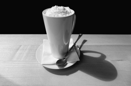 White mug on saucer with long spoon (cup, pot, tankard, pint, jar, can) stands on beige table. Hot drink coffee in cafe, day light from window. Black and white photographyの写真素材