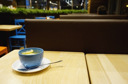 A blue half-liter mug with ginger tea and lemon stands on a light table. In the background there is an interior of a cafe with orange and blue wooden chairs.の写真素材