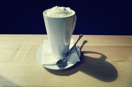 White mug on saucer with long spoon (cup, pot, tankard, pint, jar, can) stands on beige table. Hot drink coffee in cafe, day light from window. Tinted (toned) vintage imageの写真素材