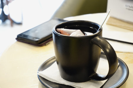 A black mug stands on a saucer with a white napkin. On the light table there is a phone behind the cup. Evening tea in the cafe.の写真素材