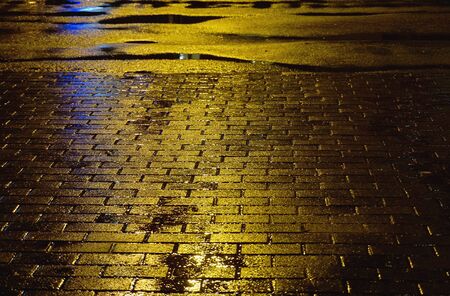 Yellow asphalt surface with puddles and paving slab. Raining weather. Night light reflected in road.