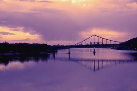 Bridge reflection in water surface of river Dnieper duaring sunset time. Toned image: purple, violet, yellow, orange colors.の写真素材