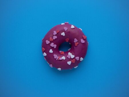 Pink donut decorated with glaze hearts isolated on a blue background top view with copy space. Sweet...