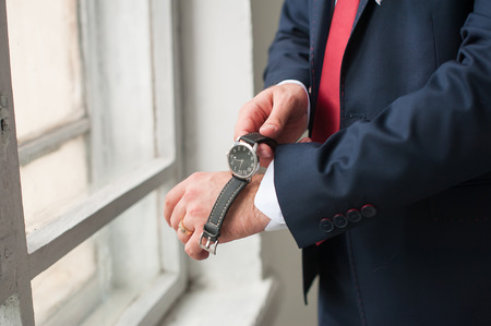 The man wears on his wrist watch while standing at the window. A man dressed in a suit, shirt and red tie. Businessman or fiance preparing himself for going out.の写真素材