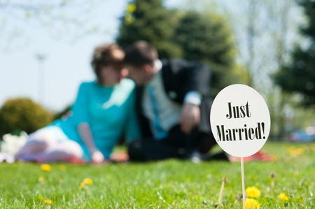 just married cardboard sign stuck in the green lawn. Couple embracing in the background. Focus on the sign.の写真素材