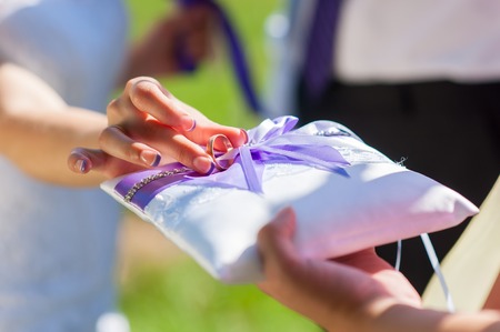 Bride gives an engagement ring to her Groom. Wedding and honeymoon concept. wedding day. outdoors. summer day. focus on hand and ring. White pillow with violet ribbonsの写真素材