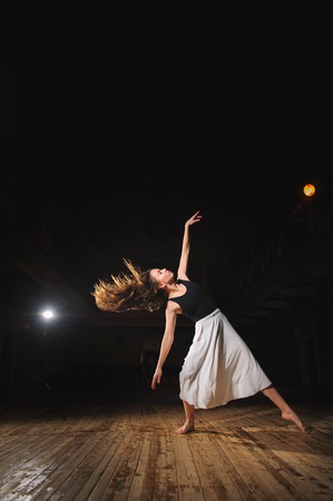 Ballerina, young brunette dancer girl in a white skirt, pointe, dance poses on stage in theater with spotlight. barefoot dancer.の写真素材